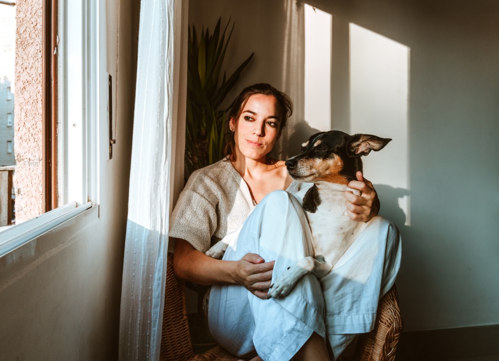 közepes felvétel egy fehér pulóveres fiatal nőről, aki egy fonott fotelben ül a lakásában hűséges kutyájával együtt