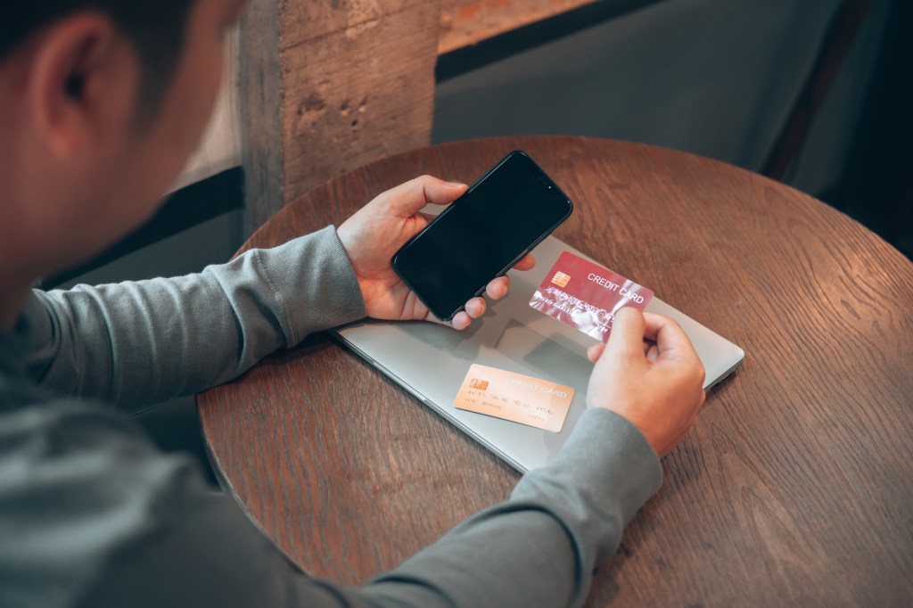 Egy férfi a kezében a telefonjával és a hitelkártyájával, hasonlóan azokhoz az emberekhez, akik online kiskutyacsalások áldozatául esnek, és megosztják a hitelkártyaadataikat.