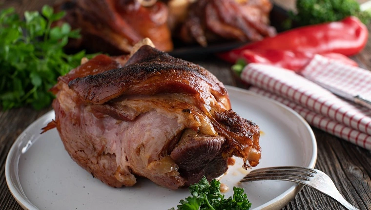 Традиционно немско месно ястие с шунка или свинско кокалче, прясно изпечено на фурна и поднесено в чиния на фона на дървена маса в селски стил. Близък план и изглед отпред