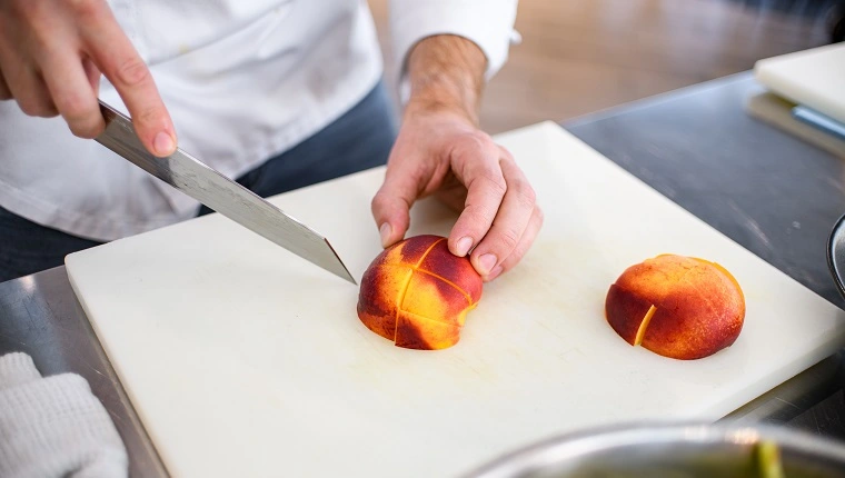 منظر من فاكهة الخوخ الناضجة شرائح على لوحة تقطيع بيضاء ويد الطهاة الذكور بسكين حاد في مكان قريب